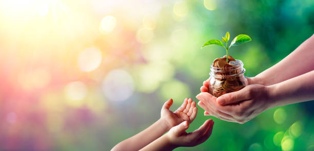 Hands of woman and child is holding coins in glass jar with young plant growing on money - Estate Planning